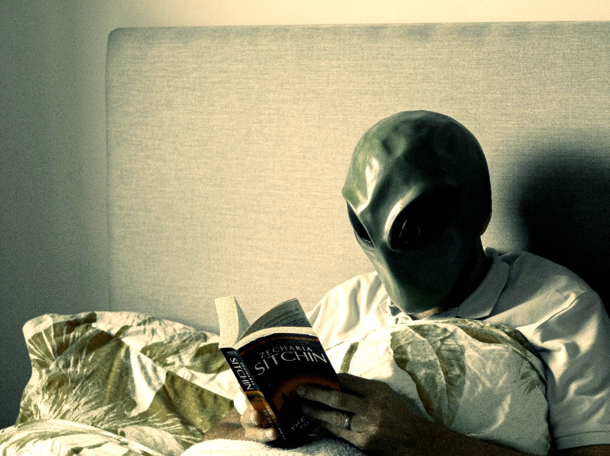 A man in an alien mask, in bed, reading a book