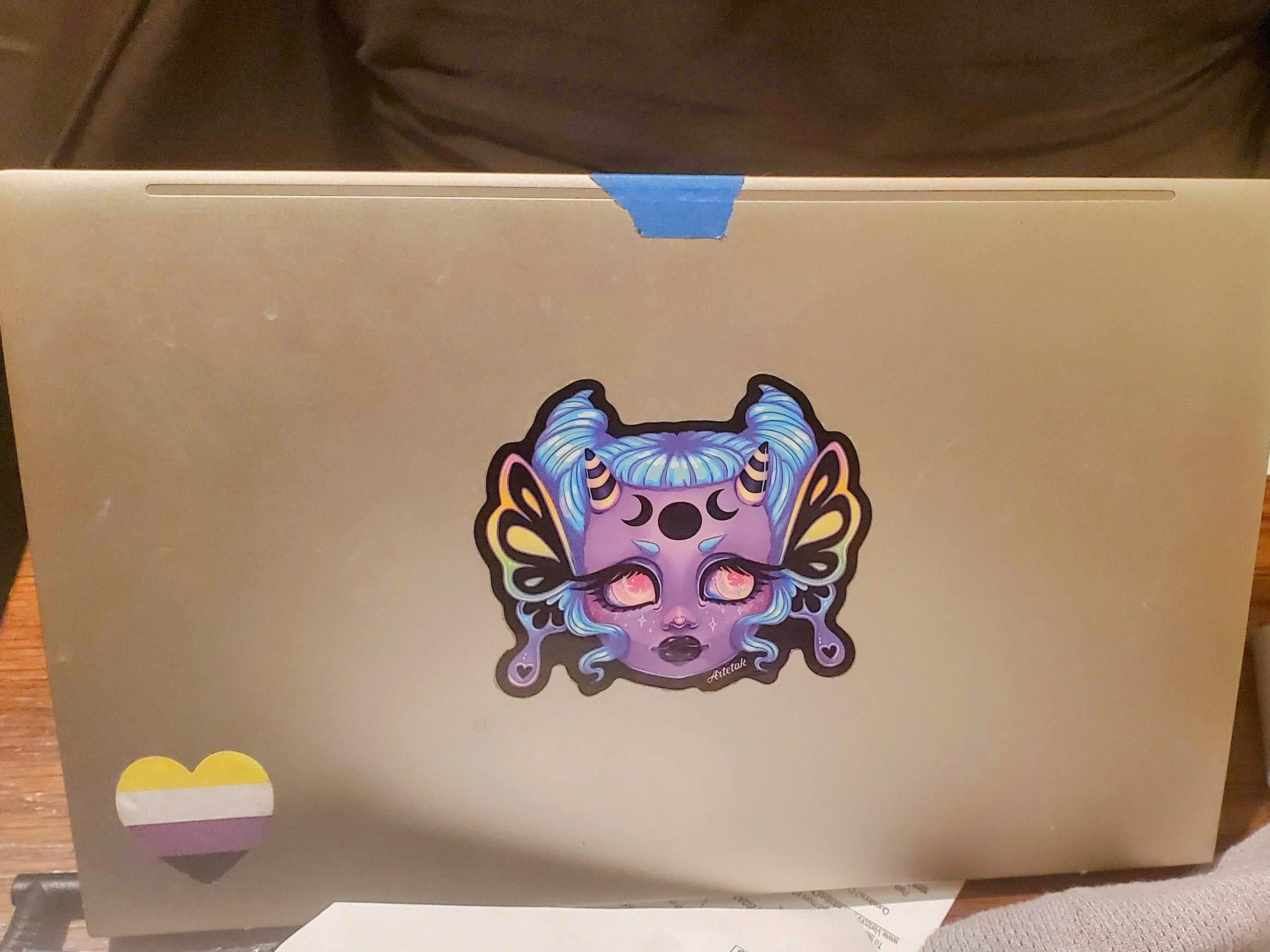 A silver laptop lid with an Artetak demon woman and a nonbinary heart sticker on it