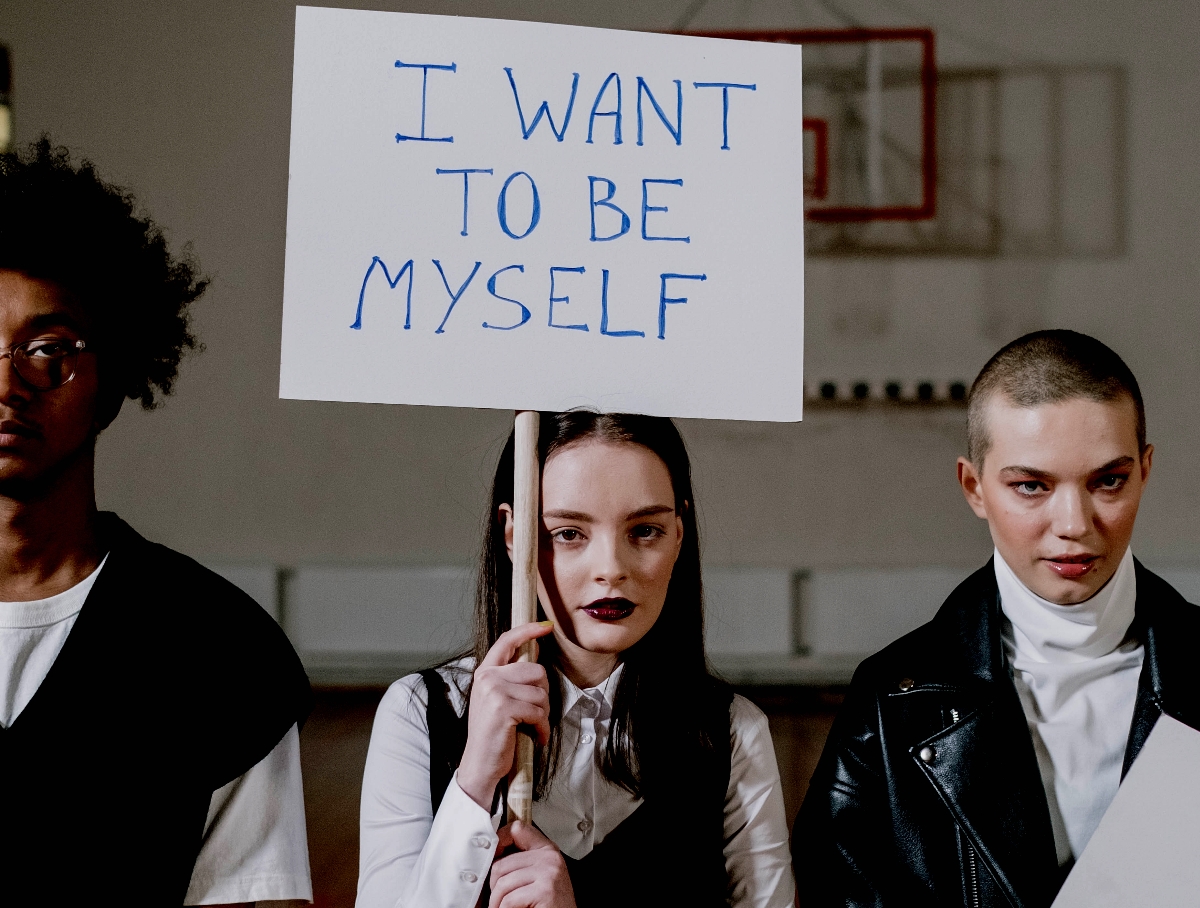 A white teenager with dark lipstick holding a sign reading I Just Want To Be Myself