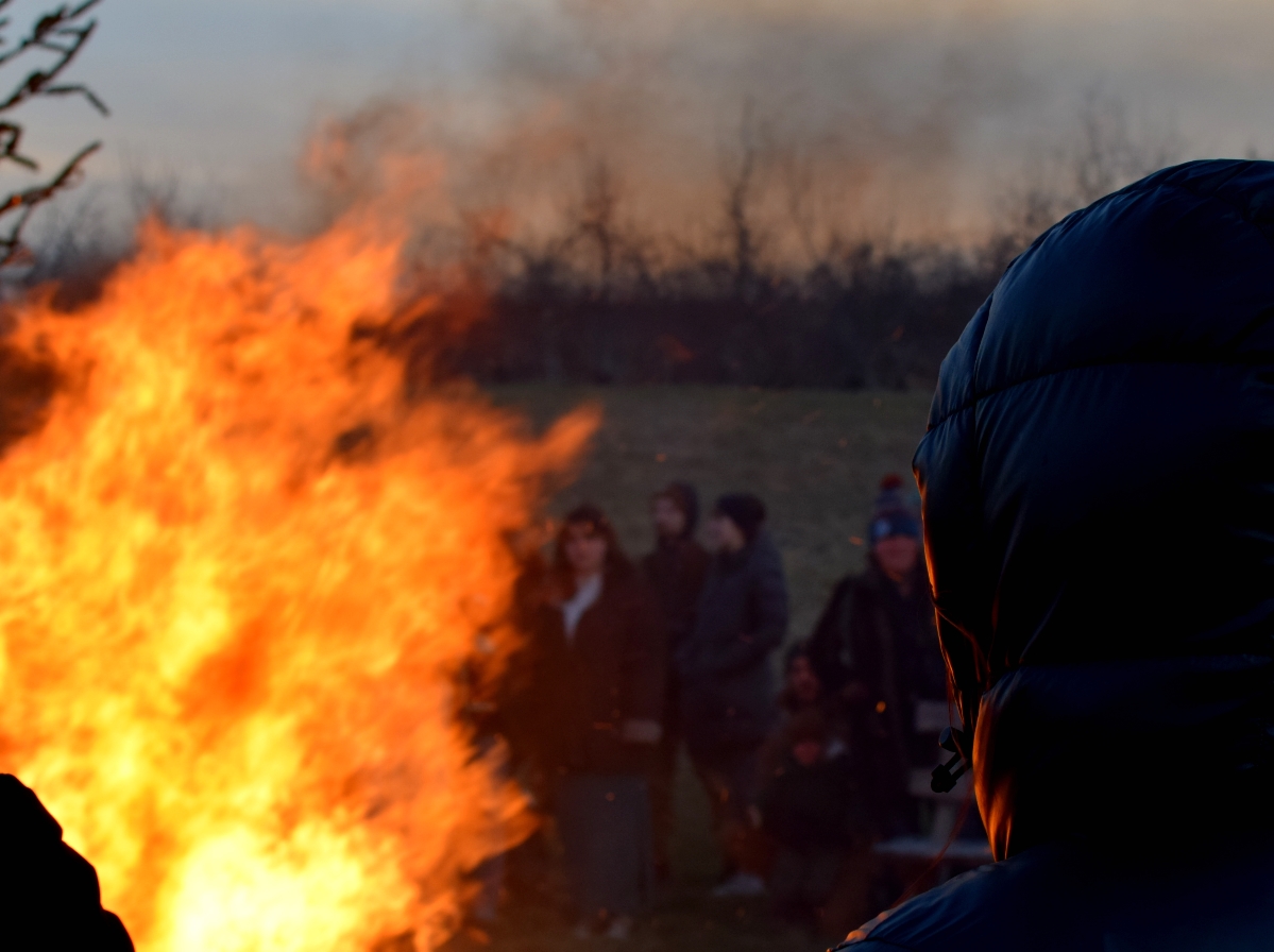 Someone in a hooded jacket watching a bonfire, the orange of it reflecting off the hood.