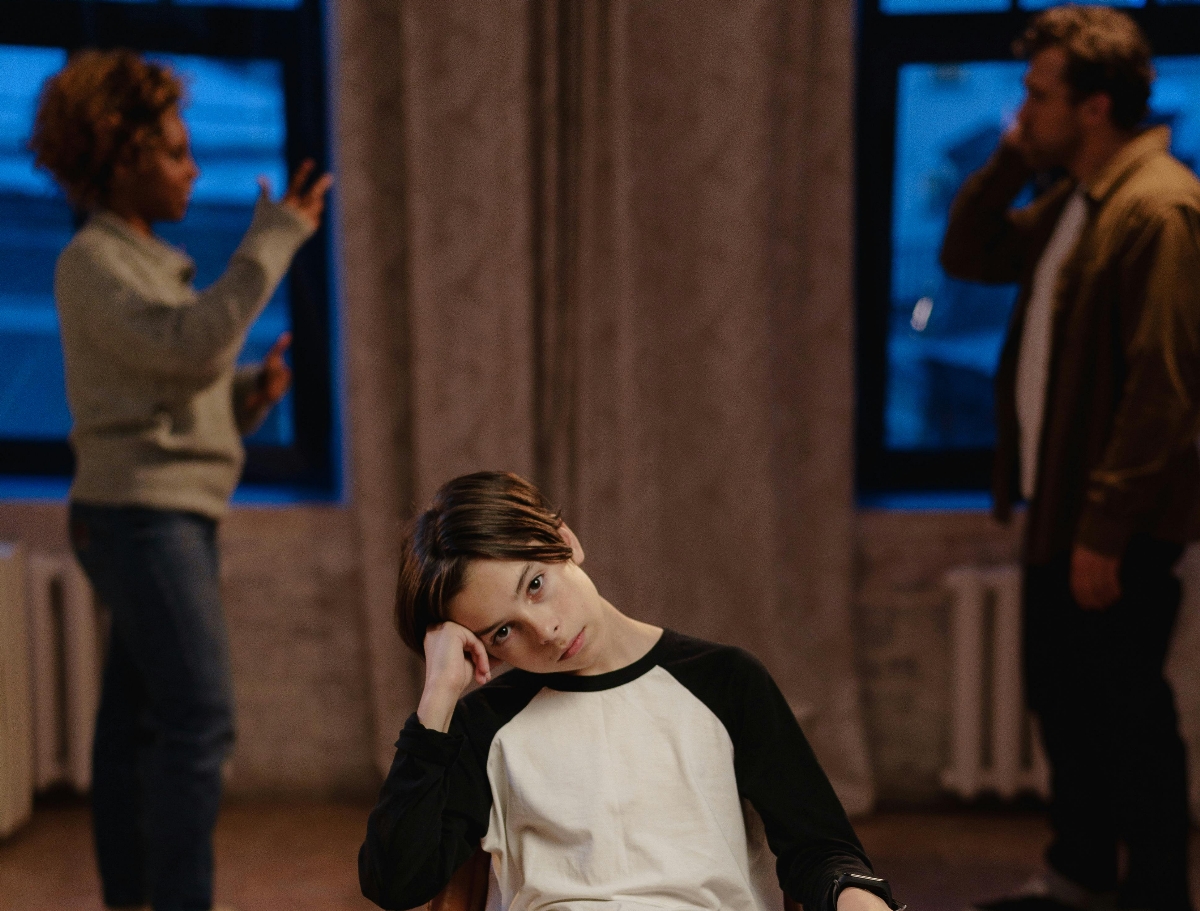 A sullen teen boy in the foreground. In the background, a Black woman with natural hair argues with a white, bearded man.
